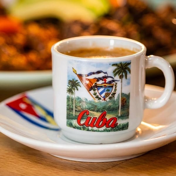 Cuban coffee in a white mug with a "Cuba" design, on a matching saucer, next to a meal.