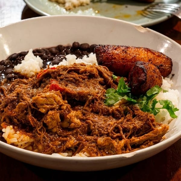 A bowl of ropa vieja with rice, black beans, and fried plantains on a wooden table.