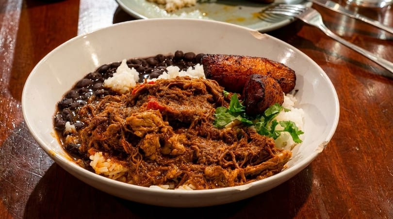 A white bowl filled with shredded meat, rice, black beans, and fried plantains on a wooden table.