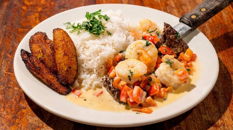 A white plate with shrimp, rice, plantains, and steak, garnished with herbs.