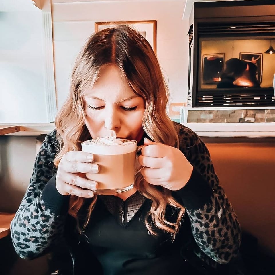 Woman in a leopard print sweater sips a frothy drink from a clear mug in a cozy restaurant.