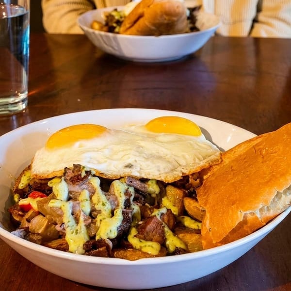 Breakfast bowl with fried eggs, potatoes, meat, green sauce, and a baguette on a wooden table.
