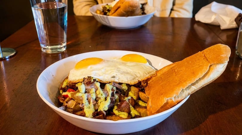 Breakfast bowl with fried eggs, potatoes, meat, green sauce, and a baguette on a wooden table.